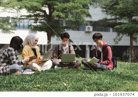 A multiethnic group of asian university students is seen studying together while sitting on the grass near the campus. 101937074
