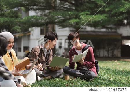 A multiethnic group of asian university students is seen studying together while sitting on the grass near the campus. 101937075