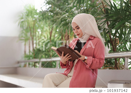 A portrait of a Muslim in white hijab university student seated inside a campus building, holding and using a laptop computer. 101937142