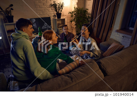 Group of happy, smiling friends sitting on couch at home in the evening and singing karaoke, talking, drinking beer and having fun Group of happy, smiling friends sitting on couch at home in the evening and singing karaoke, talking, drinking beer and having fun 101940014