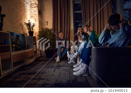 Group of friends, young people sitting on couch at home in the evening and singing karaoke, having fun, talking and drinking beer Group of friends, young people sitting on couch at home in the evening and singing karaoke, having fun, talking and drinking beer 101940015