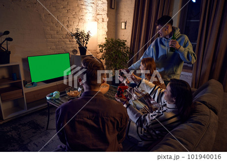 Group of friends, young men and women gathering at home in the evening, sitting on couch and playing online video games on console Group of friends, young men and women gathering at home in the evening, sitting on couch and playing online video games on console 101940016