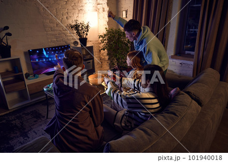 Young people, friends meeting at home in the evening, sitting on couch and playing online sport games, basketball, on console Young people, friends meeting at home in the evening, sitting on couch and playing online sport games, basketball, on console 101940018