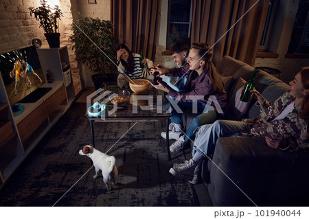 Live emotions. Friends spending time together at home in evening, sitting on couch and playing online sport games, basketball on console Live emotions. Friends spending time together at home in evening, sitting on couch and playing online sport games, basketball on console 101940044