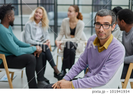 A happy young businessman, in a purple sweater, sits against the background of his colleagues A happy young businessman, in a purple sweater, sits against the background of his colleagues 101941934