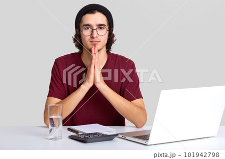 Photo of focused man with curly hair, keeps hands in praying gesture, believe in something good, wears hat and t shirt, works on modern electronic device, calculates figures, isolated over white wall 101942798