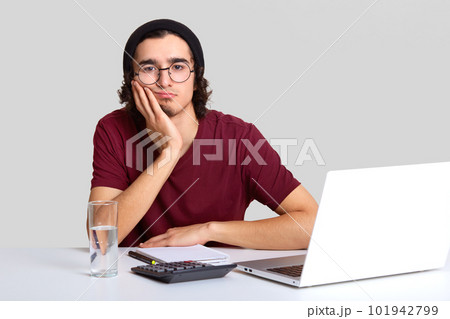 Bored male worker holds cheek with hand, looks seriously at camera, feels fatigue after working long hours, develops new project or startup, makes calculations, analyzes profits, isolated on white Bored male worker holds cheek with hand, looks seriously at camera, feels fatigue after working long hours, develops new project or startup, makes calculations, analyzes profits, isolated on white 101942799