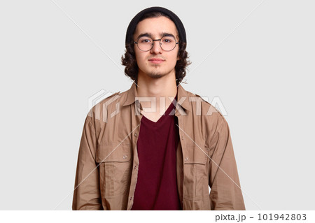 Photo of curly male teenager wears round optical spectacles, black hat, casual shirt, looks directly at camera, models against white background, being self assured. People, style, youth concept 101942803