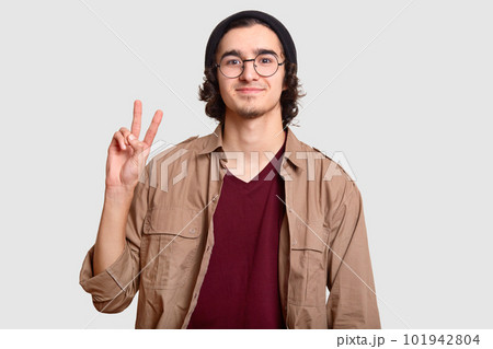 Positive curly man with little beard, makes peace sign, smiles pleasantly, dressed in fashionable hat and beige shirt, isolated over white background. Youth and body language concept. Boy says hello Positive curly man with little beard, makes peace sign, smiles pleasantly, dressed in fashionable hat and beige shirt, isolated over white background. Youth and body language concept. Boy says hello 101942804