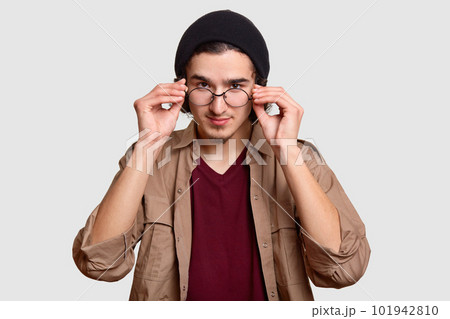 Confident handsome bearded curly man keeps hands on rim of spectacles, looks attentively, dressed in black hat and beige shirt, models against white background. People and facial expressions concept 101942810