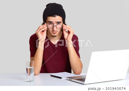 Confident student sits at workplace, looks attentively through round transparent glasses, dressed in stylish clothes, makes notes in notebook as listens webinar via laptop computer glass of water near Confident student sits at workplace, looks attentively through round transparent glasses, dressed in stylish clothes, makes notes in notebook as listens webinar via laptop computer glass of water near 101943074