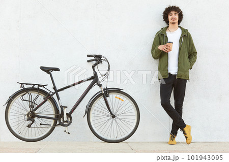Photo of attractive curly man dressed in fashionable clothes, has coffee break after riding sport bicycle, models over white concrete wall, enjoys nice trip with friend. Youth, transportation concept 101943095
