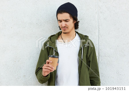 Portrait of confused unsatisfied hipster standing isolated over white wall outdoors, holding papercup with drink, undelighted with taste of drink, wearing casual clothes. Street style concept. 101943151
