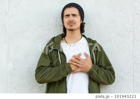 Portrait of suffering disappointed hipster putting his hands on heart, closing his eyes, heart attack, feeling pain, isolated over white wall outdoors alone. People and health problems concept. Portrait of suffering disappointed hipster putting his hands on heart, closing his eyes, heart attack, feeling pain, isolated over white wall outdoors alone. People and health problems concept. 101943221
