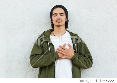 Portrait of sweet handsome youngster leaning on white wall with closed eyes, trying to feel heartbeat with two hands crossed on chest, looks concentrated, peaceful. Copy space for advertisement. 101943222