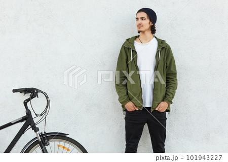 Horizontal shot of male cyclist dressed in fashionable clothes, holds hands in pockets, stands near his bicycle against white concrete wall, looks aside with thoughtful expression. Hobby concept 101943227