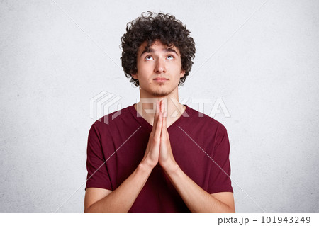 Young handsome man with curly dark hair prays with hands together. Teenager dresses casual maroon t shirt, with hope on face, looks up sky, asks for forgiveness against white wall. Religion concept. Young handsome man with curly dark hair prays with hands together. Teenager dresses casual maroon t shirt, with hope on face, looks up sky, asks for forgiveness against white wall. Religion concept. 101943249