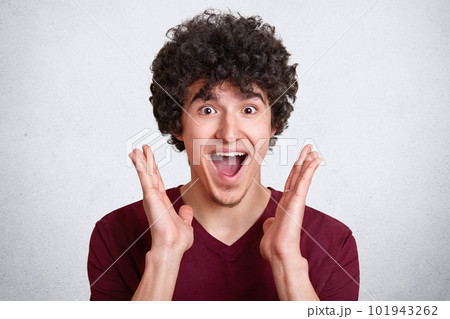 Studio shot of attractive man with dark curly hair, keeps mouth opened, screams something, stands with satisfied facial expression, has high spirit, his dreams come true, poses isolated on white wall. Studio shot of attractive man with dark curly hair, keeps mouth opened, screams something, stands with satisfied facial expression, has high spirit, his dreams come true, poses isolated on white wall. 101943262
