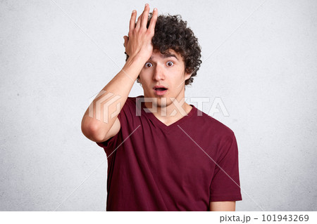 Indoor studio shot of worried curly haired young man touching his forehead with hand, covering it, opening eyes and mouth widely, looking directly at camera, wearing casual dark red t shirt. Indoor studio shot of worried curly haired young man touching his forehead with hand, covering it, opening eyes and mouth widely, looking directly at camera, wearing casual dark red t shirt. 101943269