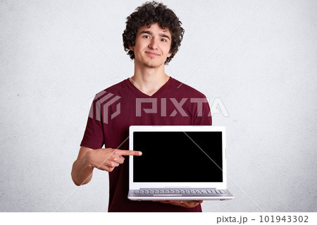 Shot of smiling handsome man wearing casual maroon t shirt standing and pointing finger on blank laptop screen over white studio background, male looking at camera with plasant facial expression. 101943302