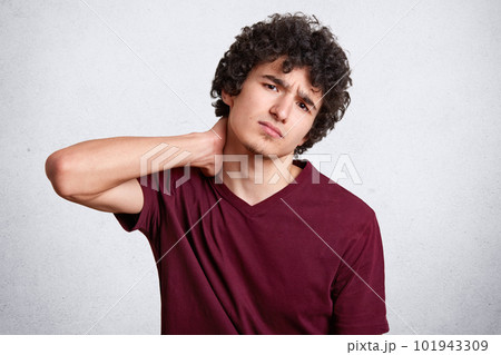 Portrait of sad handsome man with little beard and dark curly hair, wearing casual maroon t shirt, standing and holding his painful neck. Studio shot, model posing isolated over white background. Portrait of sad handsome man with little beard and dark curly hair, wearing casual maroon t shirt, standing and holding his painful neck. Studio shot, model posing isolated over white background. 101943309