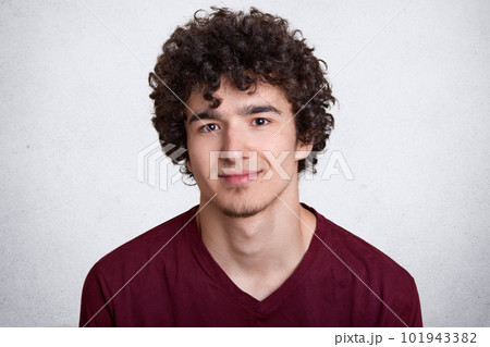 Close up portrait of sweet attractive smiling guy with curly hair and beard, having pleasant facial expression, posing isolated over white background in studio, wearing casual red sweatshirt. 101943382