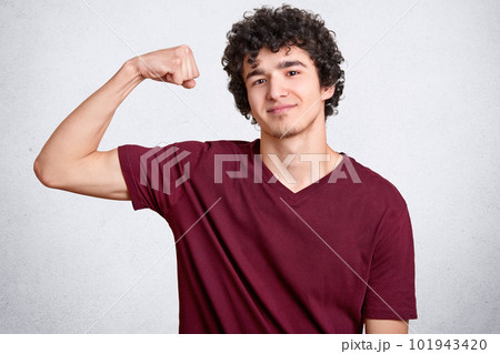 Satisfied strong man shows biceps or muscles, has delighted expression, curly hairstyle, trains every day to achieve aims and have sporty body, poses against white background, feels proud of himeself 101943420