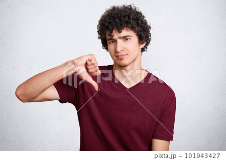 Portrait of attractive handsome male with curly hair, shows dislike gesture, keeps thumb down, wears casual t shirt, isolated over white background. Sullen European man being discontent by something 101943427