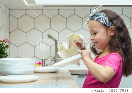 Side view of glad little girl with dark hair wearing pink T-shirt, washing dishes, drying clean white plates with cloth. 101947953