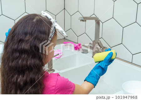 Back view of little girl wearing pink T-shirt, blue rubber gloves, standing near white sink, holding yellow sponge. Back view of little girl wearing pink T-shirt, blue rubber gloves, standing near white sink, holding yellow sponge. 101947968