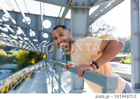 Close-up photo. Portrait of a young African American sports man standing in the stadium, doing push-ups on the bar, swinging his arms, looking at the camera smiling. 101948713