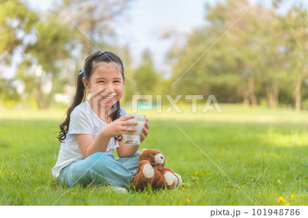 Caucasian mix girl drinking milk from glass sitting on grass at outdoor park, Whiskers from milk, Good nutrition of children concept. 101948786