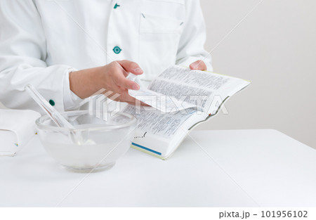 粉砕調剤とハンドブックで調べる薬剤師。乳棒と乳鉢が置かれている 101956102