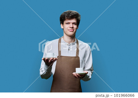 Young male waiter in apron posing on blue background with two cups of coffee Young male waiter in apron posing on blue background with two cups of coffee 101966066