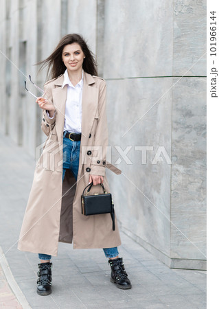 A young pretty girl in a raincoat and jeans walks around the city near a tall building, with a handbag in hand A young pretty girl in a raincoat and jeans walks around the city near a tall building, with a handbag in hand 101966144