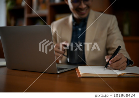 A professional adult Asian businessman taking notes on his notebook and sipping coffee 101968424