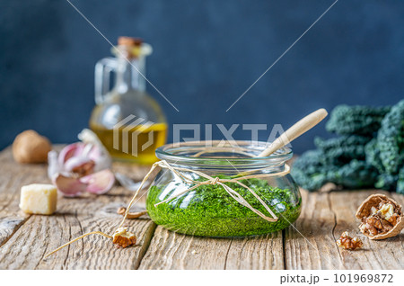 Homemade kale pesto served in a glass jar on wooden surface with dark background and basic ingredients - kale, walnuts, parmesan, garlic and olive oil 101969872