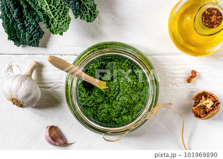 Homemade kale pesto sauce served in a glass bowl over white wooden surface with basic ingredients - kale leaves, walnuts, parmesan, garlic and olive oil Homemade kale pesto sauce served in a glass bowl over white wooden surface with basic ingredients - kale leaves, walnuts, parmesan, garlic and olive oil 101969890
