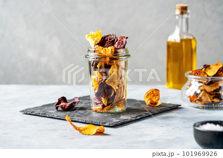 Healthy vegetable chips in a glass jar on stone cutting board with marine salt and oil aside. Concrete background Healthy vegetable chips in a glass jar on stone cutting board with marine salt and oil aside. Concrete background 101969926