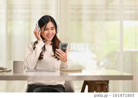 Cheerful asian woman listening to music in wireless headphones and using smartphone at desk in living room 101971805