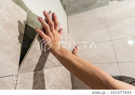 Close-up of worker tiler hands installing light beige ceramic tiles on walls of future bathroom. Tiles installation, home improvement, renovation and construction, comfortable modern home concept. Close-up of worker tiler hands installing light beige ceramic tiles on walls of future bathroom. Tiles installation, home improvement, renovation and construction, comfortable modern home concept. 101977693