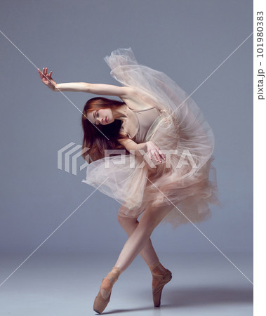 Sensual red haired ballerina wearing tulle dress dancing with emotions over grey studio background. Contemporary dance. Weightless easy jump 101980383