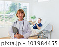 the doctor is examining the patient in the hospital. Caucasian female doctor in a gown Hand holding tablet looking at camera with patient in background in hospital. 101984455