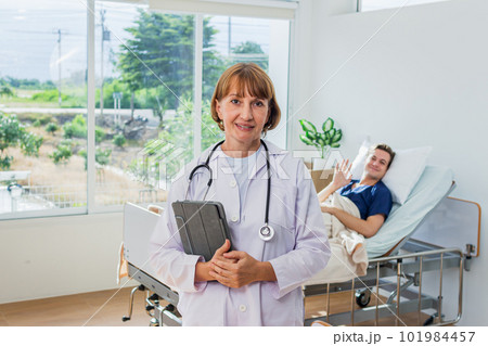 the doctor is examining the patient in the hospital. Caucasian female doctor in a gown Hand holding tablet looking at camera with patient in background in hospital. 101984457