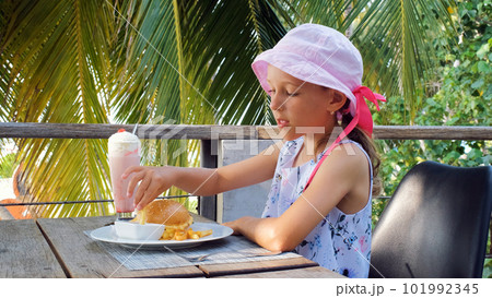 Little girl eating french fries and cheeseburger while sitting on a terrace in tropical island Little girl eating french fries and cheeseburger while sitting on a terrace in tropical island 101992345