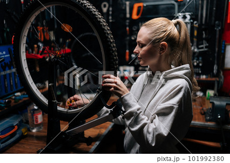 Portrait of pretty blonde female cycling repairman checking bicycle wheel spoke with bike spoke key in repair workshop with dark interior. Concept of professional maintenance of bicycle transport. Portrait of pretty blonde female cycling repairman checking bicycle wheel spoke with bike spoke key in repair workshop with dark interior. Concept of professional maintenance of bicycle transport. 101992380