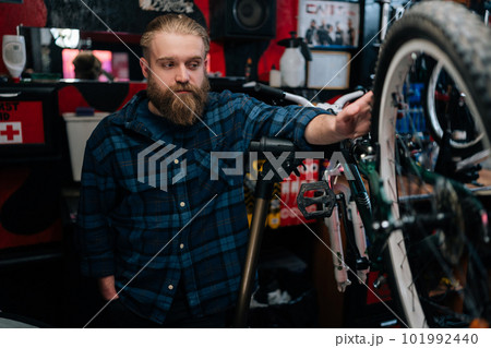 Portrait of pensive bearded cycling mechanic male standing by bicycle in repair bike workshop with dark interior, thoughtful looking away. Concept of professional repair and maintenance of bicycle. Portrait of pensive bearded cycling mechanic male standing by bicycle in repair bike workshop with dark interior, thoughtful looking away. Concept of professional repair and maintenance of bicycle. 101992440