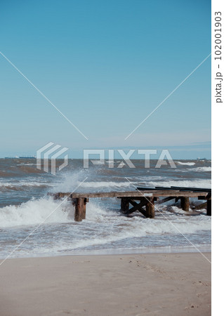 Storm sea waves empty wooden pier in dramatic blue sky background. Seascape with running splashing sea waves. Sandy beach vacation getaway in Gdansk Poland.  102001903