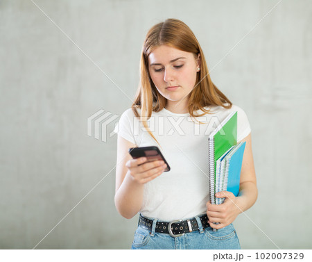Positive teenage girl, student in casual clothes using mobile phone and holding spiral notebooks against gray wall background, copy space for mock up Positive teenage girl, student in casual clothes using mobile phone and holding spiral notebooks against gray wall background, copy space for mock up 102007329