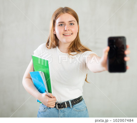 Smiling teenage girl, student with notebooks under her arm and mobile phone with blank black screen  102007484